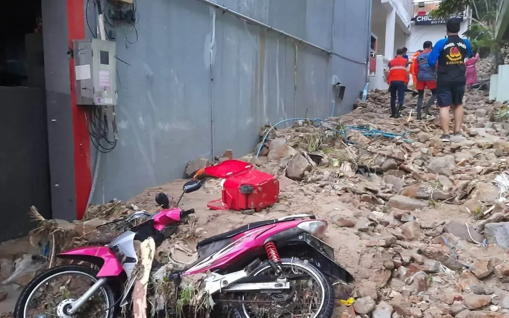 People walk through a rocky flood-damaged lane beside a fallen motorbike.