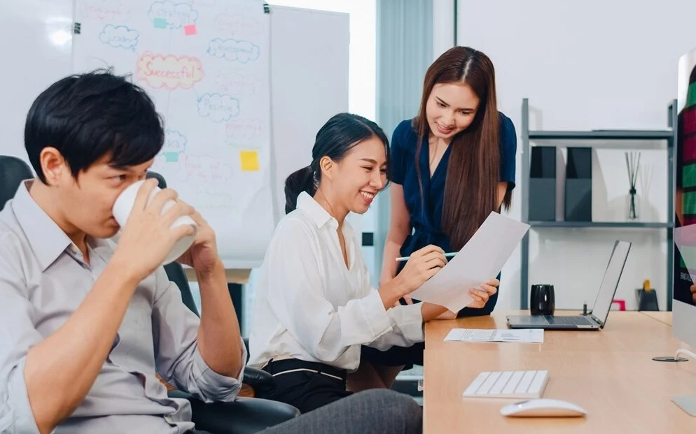 Coworkers discuss papers beside a computer in an office.