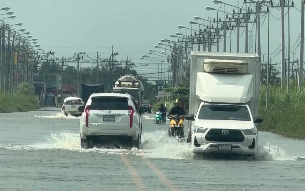 Vehicles and motorcyclists drive through a flooded road beneath rows of utility poles.