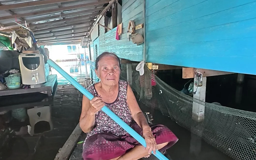 An elderly woman sits in a flooded home holding a blue pole.