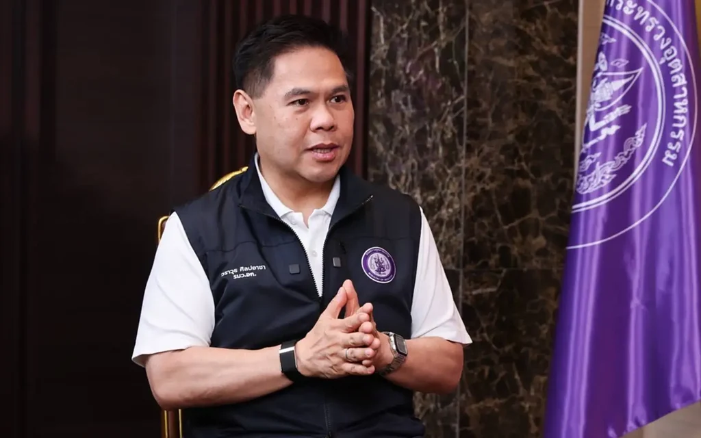 A government official speaks beside a purple flag in an indoor setting.
