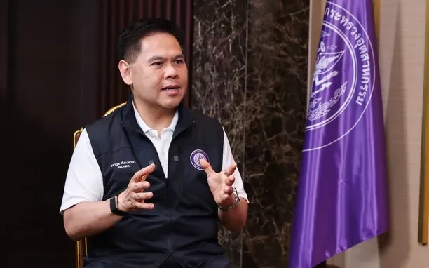 A minister speaks beside a purple agency flag during an interview.