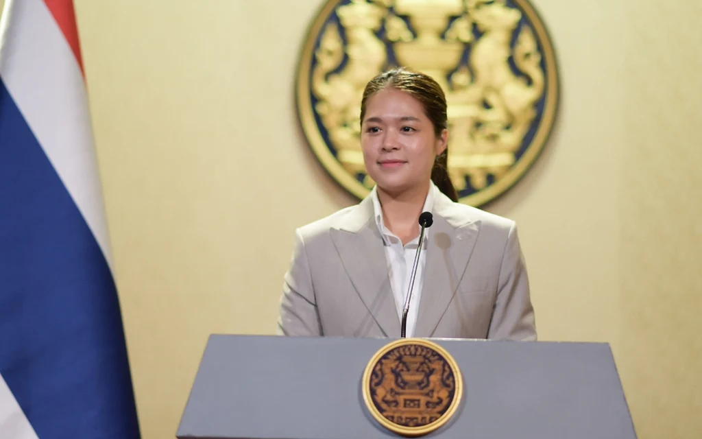 A government spokesperson speaking at a podium with the Thai flag.