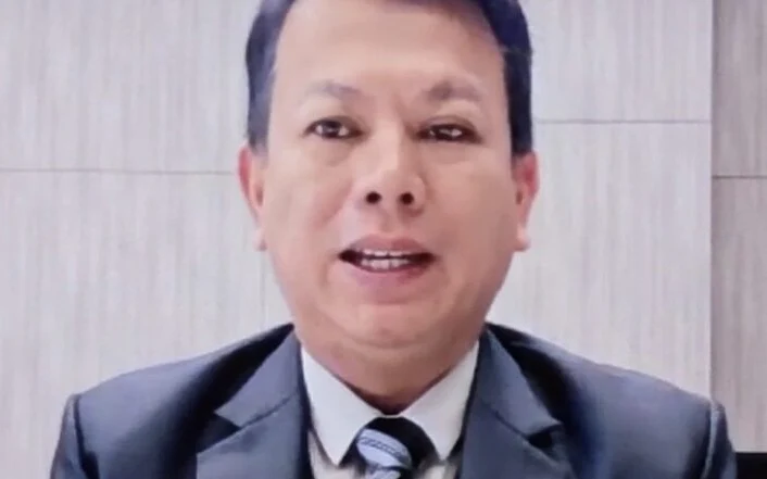 Man in a dark suit speaking to the camera indoors