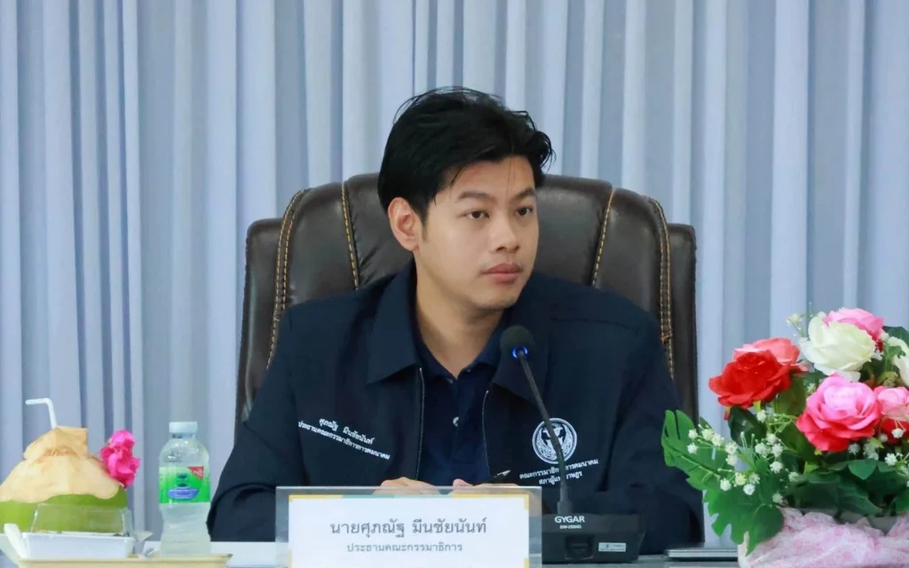 A man speaks at a meeting, seated behind a nameplate and flowers.